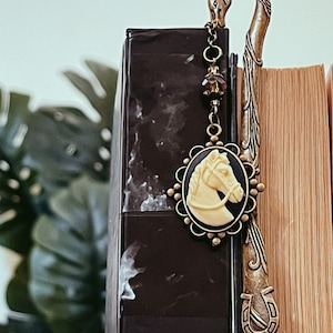 May include: A bronze-toned metal bookmark with a horse head design. The bookmark features a cameo with a cream-colored horse profile on a black background. The design includes a horseshoe and decorative chain.