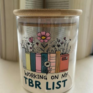 May include: A clear glass jar with a bamboo lid, featuring a colorful illustration of books and flowers. The text "WORKING ON MY TBR LIST" is printed on the jar. The background includes open books.