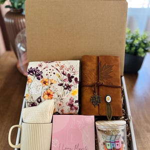 May include: A gift box containing a floral fabric, a brown leather journal, a cream mug, a pink box with text, and a candle. The candle has a wooden lid and a label reading "TBR QUEEN". The box is made of cardboard.