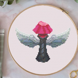 May include: Cross-stitch embroidery of a winged torch with a pink flame and grey wings, set in a wooden embroidery hoop. The design is on a white fabric background. The craft project is displayed on a pink surface.