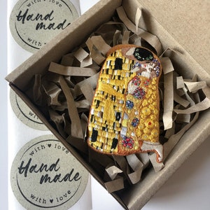 May include: A small, embroidered patch of Gustav Klimt's The Kiss artwork in a cardboard box with brown paper packing material. The patch is yellow, gold, and black with small details in red, blue, and white. The box has a sticker that says "with love handmade with love".