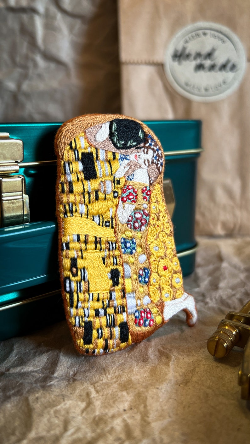 May include: A hand-stitched embroidered patch depicting Gustav Klimt's "The Kiss" painting. The patch features a yellow, gold, and black color scheme with intricate details.