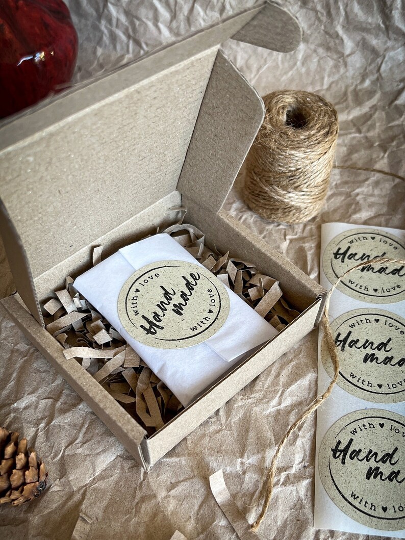 May include: A small cardboard box with a white tissue paper inside. The box is filled with brown crinkled paper. The box has a sticker on it that says "Hand made with love".
