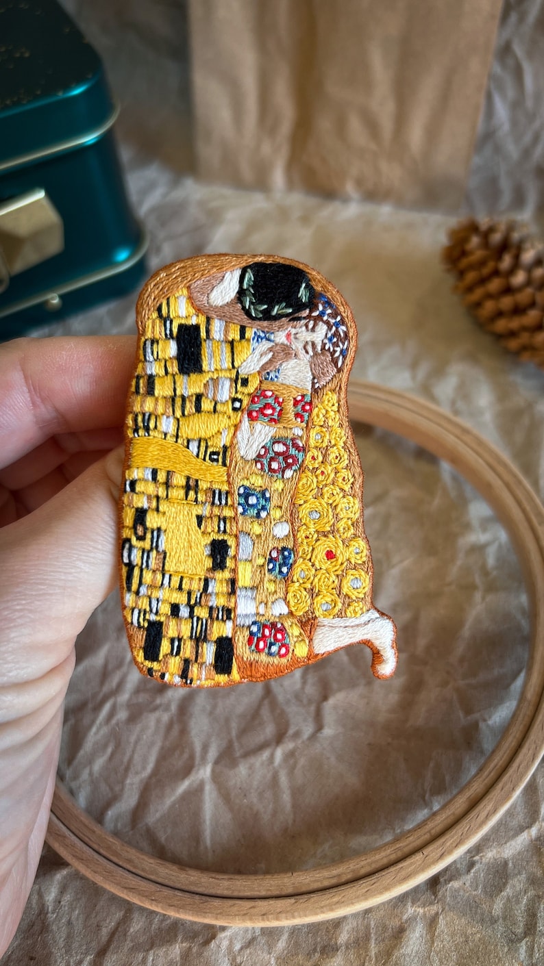 May include: Embroidered artwork of Gustav Klimt's The Kiss, featuring a couple embracing in a mosaic of yellow, white, and black squares. The artwork is framed in a wooden hoop.