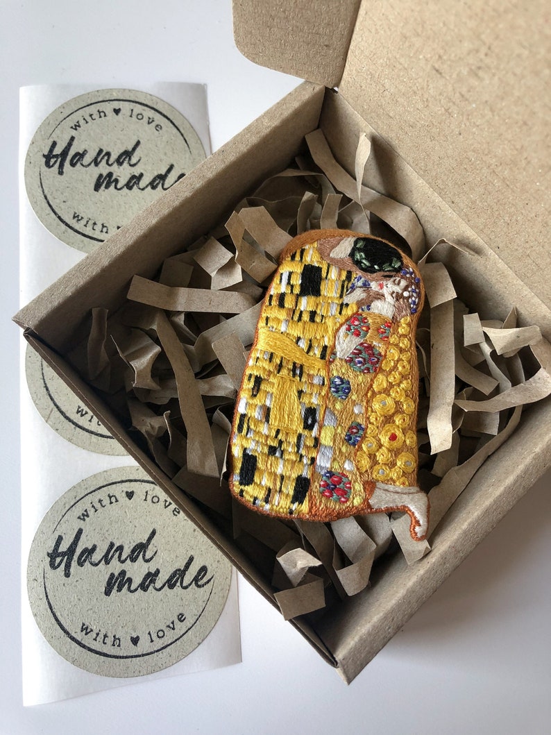 May include: A small, embroidered patch of Gustav Klimt's The Kiss artwork in a cardboard box with brown paper packing material. The patch is yellow, gold, and black with small details in red, blue, and white. The box has a sticker that says "with love handmade with love".