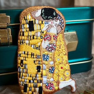 May include: A close-up of a hand-stitched embroidered patch depicting Gustav Klimt's The Kiss painting. The patch features the iconic couple in a warm embrace, with the man's robe adorned with a yellow and black checkered pattern and the woman's dress in a mosaic of colorful flowers and geometric shapes.