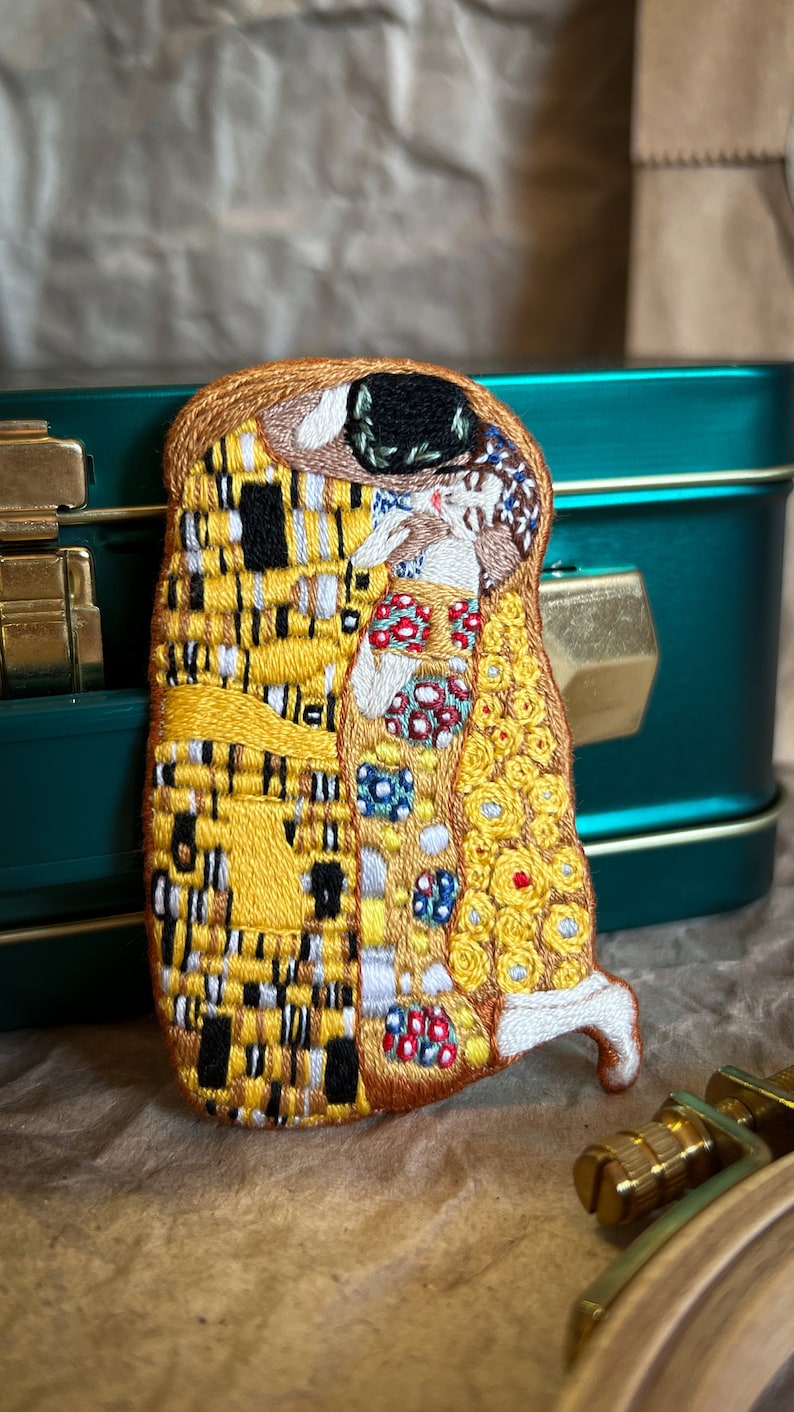 May include: A close-up of a hand-stitched embroidered patch depicting Gustav Klimt's The Kiss painting. The patch features the iconic couple in a warm embrace, with the man's robe adorned with a yellow and black checkered pattern and the woman's dress in a mosaic of colorful flowers and geometric shapes.