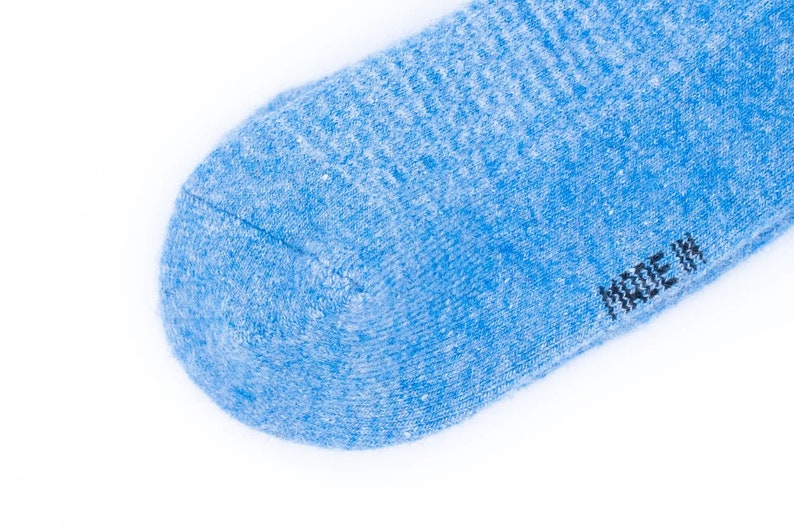 Puede incluir: Primer plano de un calcet&iacute;n azul claro con un patr&oacute;n de punto texturizado. El calcet&iacute;n tiene una puntera redondeada y el texto "MADE IN" es visible en negro. El fondo es blanco.