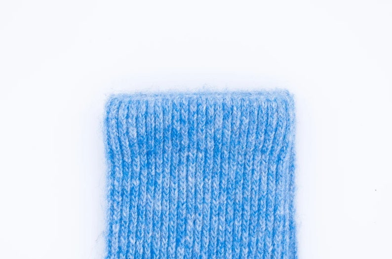 Puede incluir: Primer plano del pu&ntilde;o de un calcet&iacute;n de punto azul claro. El pu&ntilde;o es acanalado y tiene un aspecto suave y texturizado. El calcet&iacute;n est&aacute; hecho de un hilo azul claro con motas blancas, creando un efecto jaspeado. El fondo es blanco liso.
