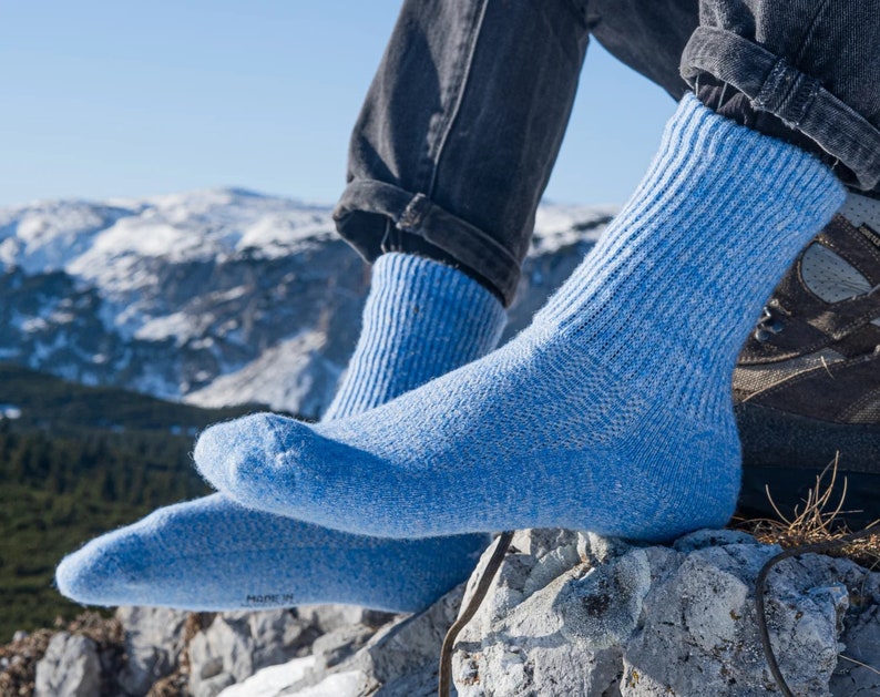 Puede incluir: Calcetines azul claro, con textura acanalada, se usan con vaqueros oscuros. Los calcetines descansan sobre una superficie rocosa, con una cordillera nevada al fondo. Los calcetines parecen estar hechos de un material grueso y c&aacute;lido.