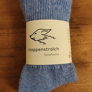 Puede incluir: Un par de calcetines de lana azul claro, doblados y empaquetados. Los calcetines est&aacute;n envueltos con una etiqueta blanca con una ilustraci&oacute;n de oveja y el texto "steppenstrolch Schafwolle".