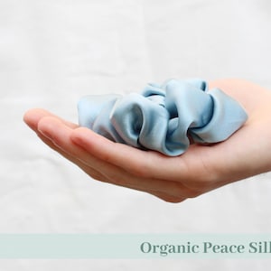 May include: A light blue scrunchie made of organic peace silk. The scrunchie is held in an open hand against a white background. The text "cruelty-free" and "Organic Peace Silk" are visible.