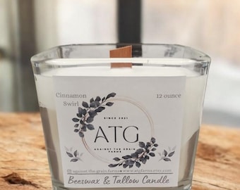 May include: A clear glass square candle holder with a wooden wick, filled with white wax. The label reads "Cinnamon Swirl" and "12 ounce". The candle is labeled "ATG Against The Grain Farms" and "Beeswax & Tallow Candle".