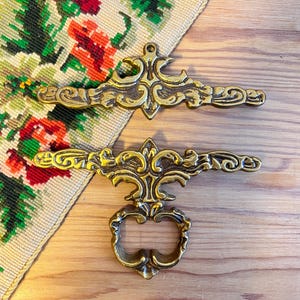 May include: Two ornate brass furniture pulls with intricate scrollwork and raised relief detailing. One pull features a horizontal bar design, while the other includes a matching horizontal bar with an attached circular drop handle for gripping.