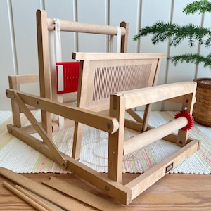 Op de afbeelding: Een tafelweefgetouw van lichtgekleurd hout met een stijf riet met verticale sleuven, een rode plastic spoel en een rood spanwiel. Het weefgetouw bevat diverse houten weefstokken en is opgebouwd met een stevig frame en metalen onderdelen.