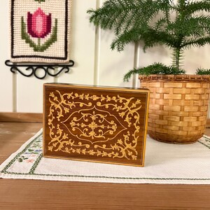 May include: A wooden box with a decorative inlay of a floral pattern in gold. The box is closed and sitting on a white tablecloth.
