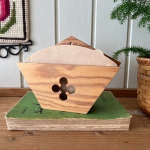May include: A wooden coffee filter holder with a decorative design. The holder is made of light brown wood and has a four-leaf clover design cut out of the front. The holder is sitting on top of a stack of books.