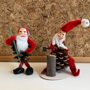 May include: Two red and white Santa Claus figures made of pipe cleaners and pine cones. One Santa is holding a green stick and the other is holding a small axe and is sitting on a log.