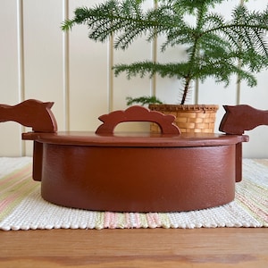 May include: A round, reddish-brown wooden box with a lid featuring a central handle and two decorative horse head silhouettes on opposite sides. The container has a smooth, painted finish and a simple, functional design for storage purposes.