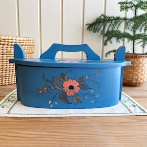 Peut inclure: Une boîte en bois bleue avec une poignée et un motif floral. La boîte est peinte avec un motif floral dans des tons orange, gris et blanc. Elle repose sur un tissu brodé blanc et vert.