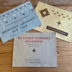 Peut inclure: Trois livres anciens sur une surface en bois. Les livres présentent des titres en français, danois et suédois, avec des illustrations de motifs de broderie. Les couvertures des livres sont dans des tons de bleu, beige et blanc cassé, avec du texte en noir et rouge.