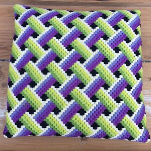 Könnte beinhalten: Ein quadratisches Kissen mit einem geometrischen Muster in Grün-, Lila-, Schwarz- und Weißtönen. Das Kissen ist aus einem weichen, strukturierten Stoff gefertigt.