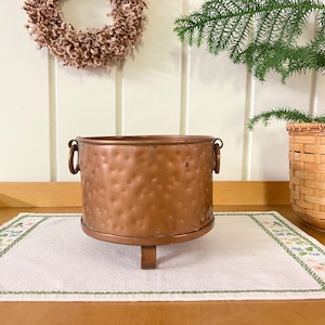 May include: A hammered copper planter with three legs. The planter is on a white embroidered table runner with a green border.