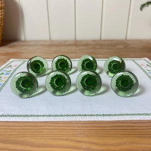 以下が含まれることがあります： 白い刺繍布の上に、花の模様が入った緑色のガラス製キャビネットノブが8つ並んでいます。ノブは丸く、濃い緑色の中心部があります。ノブは木製の表面にあります。