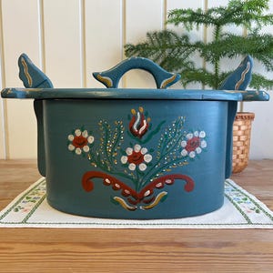 Op de afbeelding: Een blauwgroene houten container met deksel, voorzien van een handgeschilderd bloemmotief in rood, wit en groen. Het deksel heeft een centrale handgreep en twee zijhandgrepen met gouden accenten. De container heeft een matte afwerking en ronde vorm.
