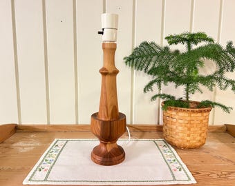 May include: A wooden table lamp with a white lampshade. The lamp has a unique, turned wood base and a natural wood finish. The lamp is sitting on a white table runner with a floral pattern.
