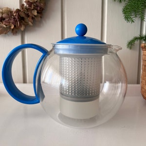 May include: A clear glass teapot with a blue lid and handle. The teapot has a built-in strainer for loose leaf tea.