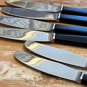 Könnte beinhalten: Fünf silberne Buttermesser mit schwarzen Kunststoffgriffen. Die Messer sind in einer Reihe angeordnet, wobei die Griffe nach oben zeigen. Die Messer sind alle gleich groß und gleich geformt.