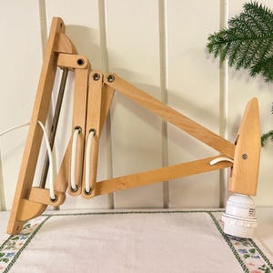 May include: A wooden wall-mounted articulated lamp with a natural finish. The lamp features a concertina-style arm and a white bulb socket. The design includes a white power cord and is mounted on a wooden base. The lamp is approximately 30 inches long when fully extended.