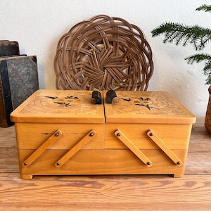 May include: A light-colored wooden sewing box with a cantilevered accordion-style opening mechanism. The top lids feature dark, inlaid floral motifs and two black handles. The box has a rectangular base and is constructed from smooth, polished wood.