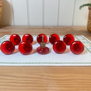 May include: Ten red glass spheres, including a bottle stopper, are displayed on a white embroidered cloth. The spheres are evenly spaced, highlighting their round shape and glossy finish. The background features a wooden surface and a wicker basket.