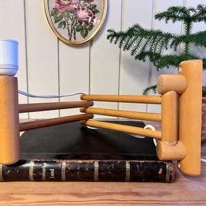May include: Wooden desk lamp with a light bulb, featuring a unique design with horizontal wooden rods. The lamp is placed on a vintage book. The lamp is made of light and dark brown wood.