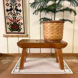 Puede incluir: Un taburete de madera con un acabado natural se encuentra sobre una superficie de madera con un encaje blanco. Una cesta de mimbre llena de una planta verde se encuentra encima del taburete.