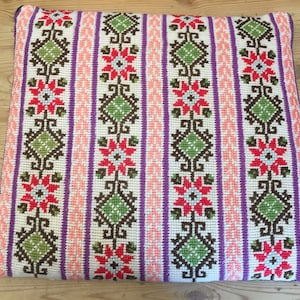 Könnte beinhalten: Ein weißes Kissen mit einem bunten Stickmuster. Das Muster zeigt sich in sich wiederholenden Mustern von Blumen, Rauten und Spiralen in den Farben Rosa, Grün, Braun und Lila.