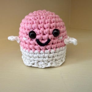May include: A handmade crochet creature with a pink head and a white base. It has black button eyes, a black stitched smile, and small white arms. The toy is designed to look like a friendly character.