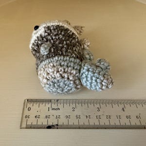 以下が含まれることがあります： グレー、ブラウン、白の色合いの手作りの編みぐるみアライグマのおもちゃ。おもちゃは横に寝ており、サイズの目安として定規が添えられています。おもちゃの長さは約7.6cmです。