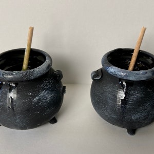 May include: Two small, black cauldron-shaped pots with white accents around the rim and on the body. Each pot has a wooden stick inside. The pots have three small legs and two handles on the sides. The pots are likely for decorative purposes.