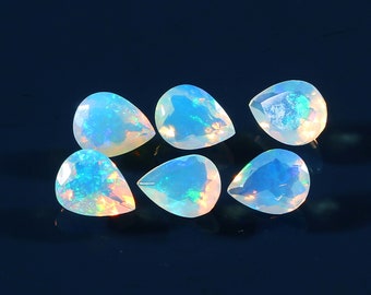May include: Six pear-shaped opals with a shimmering, iridescent play of color. The gemstones display a milky white base with flashes of blue, green, and orange. The opals are arranged on a dark blue surface.