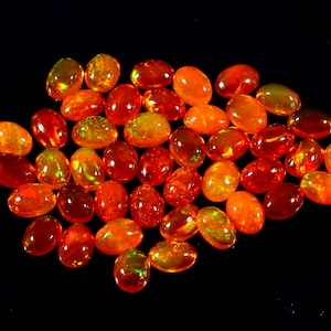 Könnte beinhalten: Eine Sammlung ovaler, orangefarbener und gelber Edelsteine mit einem feurigen, irisierenden Schimmer. Die Steine sind lose auf einem schwarzen Hintergrund angeordnet.