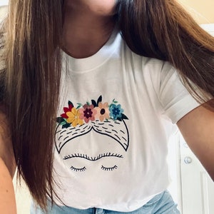 Pu&ograve; includere: Maglietta bianca con un'immagine ricamata di Frida Kahlo con una corona di fiori. L'immagine &egrave; in bianco e nero con fiori colorati.