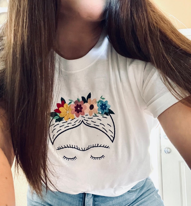 Pu&ograve; includere: Maglietta bianca con un'immagine ricamata di Frida Kahlo con una corona di fiori. L'immagine &egrave; in bianco e nero con fiori colorati.