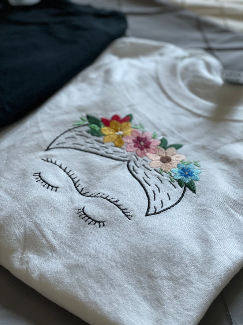 Pu&ograve; includere: Maglietta bianca con un ricamo che rappresenta il viso di Frida Kahlo con una corona di fiori. I fiori sono rosa, gialli, blu e rossi.