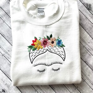 Pu&ograve; includere: Felpa bianca con un'immagine ricamata di Frida Kahlo con una corona di fiori. I fiori sono rossi, gialli, rosa e blu.