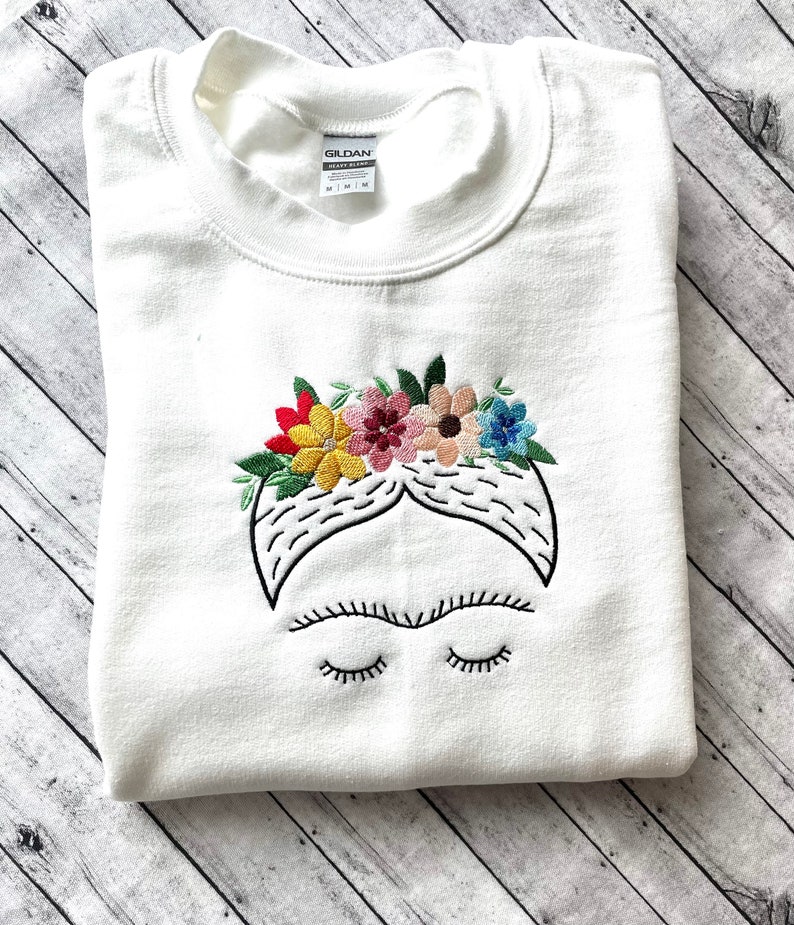Pu&ograve; includere: Felpa bianca con un'immagine ricamata di Frida Kahlo con una corona di fiori. I fiori sono rossi, gialli, rosa e blu.