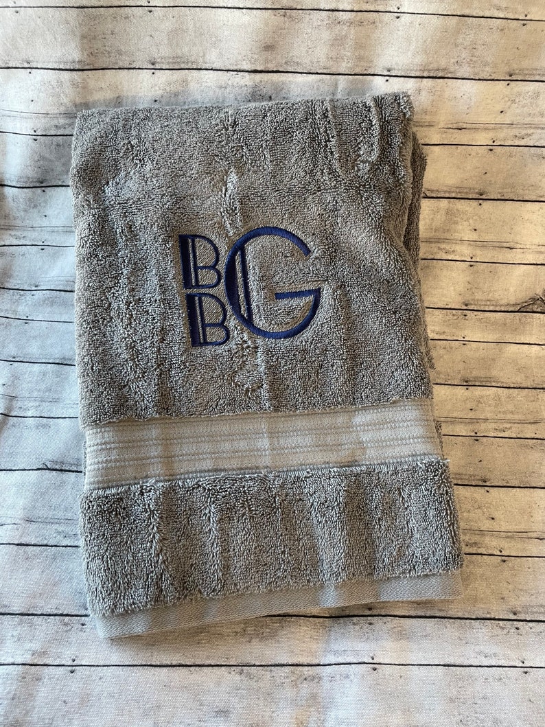 May include: A gray bath towel with a white stripe and the letters "BG" embroidered in navy blue.