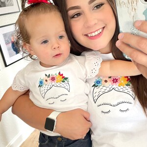 Pu&ograve; includere: Una donna e una bambina piccola indossano magliette bianche abbinate con un'immagine ricamata di Frida Kahlo. La bambina indossa jeans blu e la donna tiene la bambina. La donna indossa una maglietta bianca con bordi in pizzo.